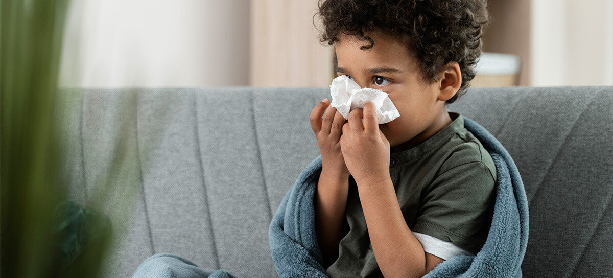 Little boy sitting on couch with blanket while blowing nose.